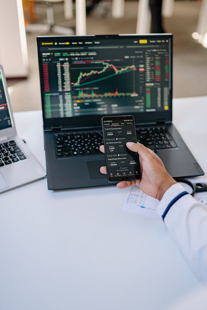 Personne analysant des données boursières sur un ordinateur portable et un smartphone à un bureau.